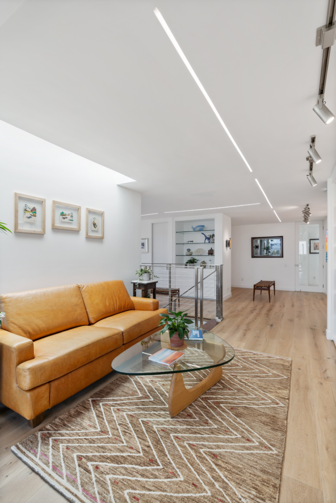 Upper-level sitting area with tan leather sofa, glass coffee table, wood flooring, cable railing and linear ceiling lights.