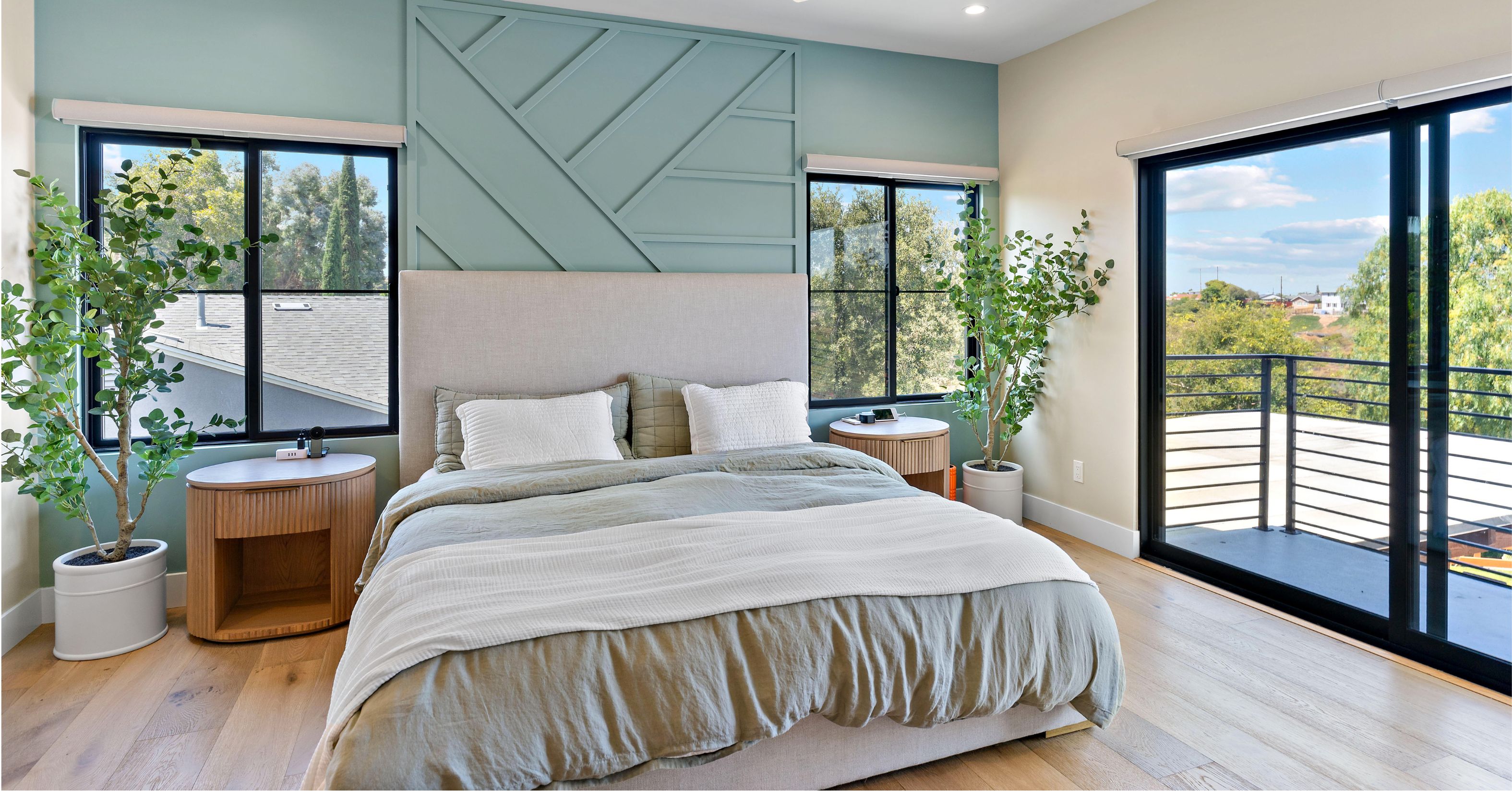 Primary bedroom with upholstered bed, green accent wall, black-framed windows, wood flooring, and private balcony.
