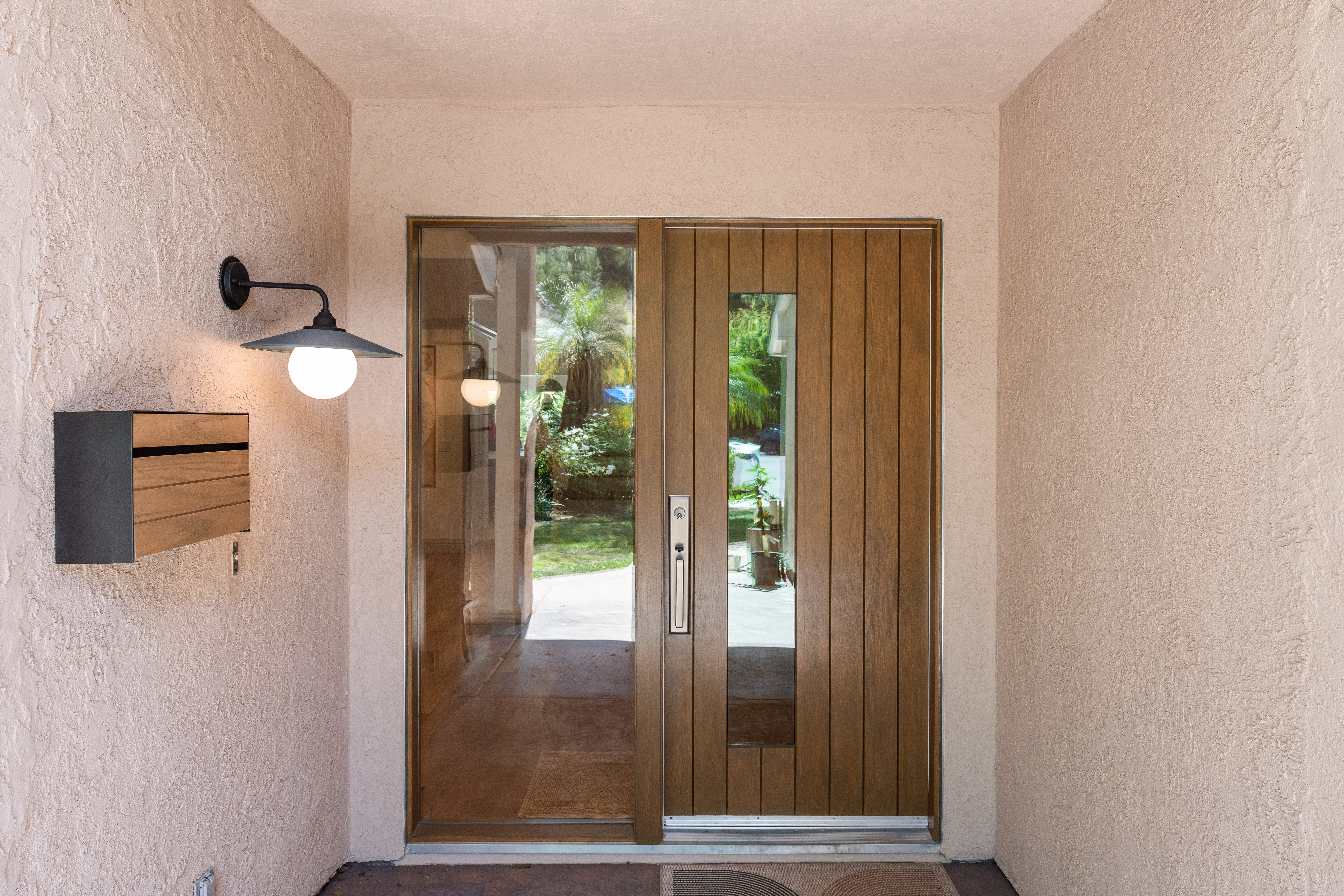 Wood front door with narrow glass panel and sidelight, textured stucco walls, black sconce and wood mailbox.