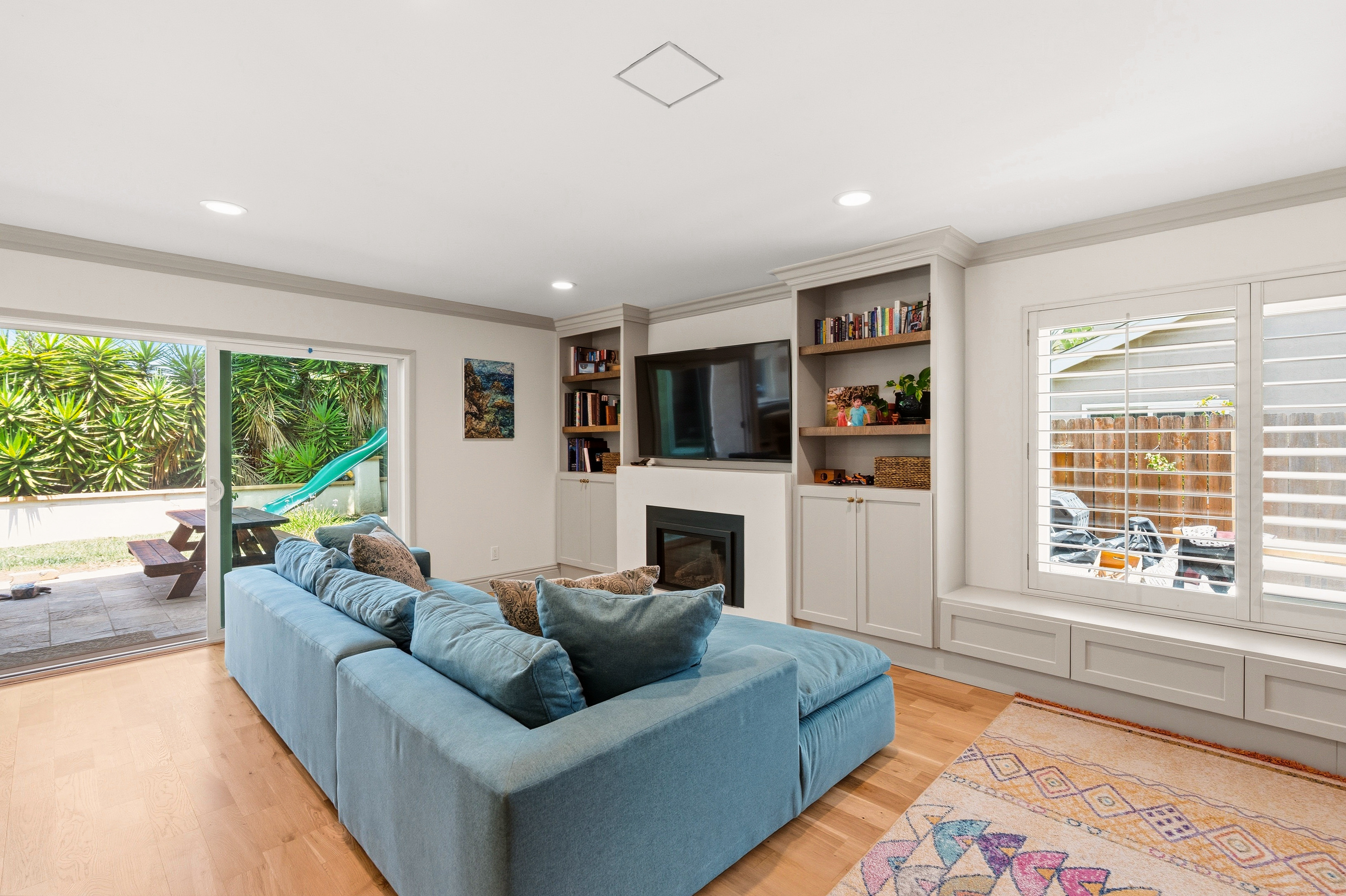 Family room with blue sectional, built-in media wall, fireplace, open shelving, window seat and backyard doors.