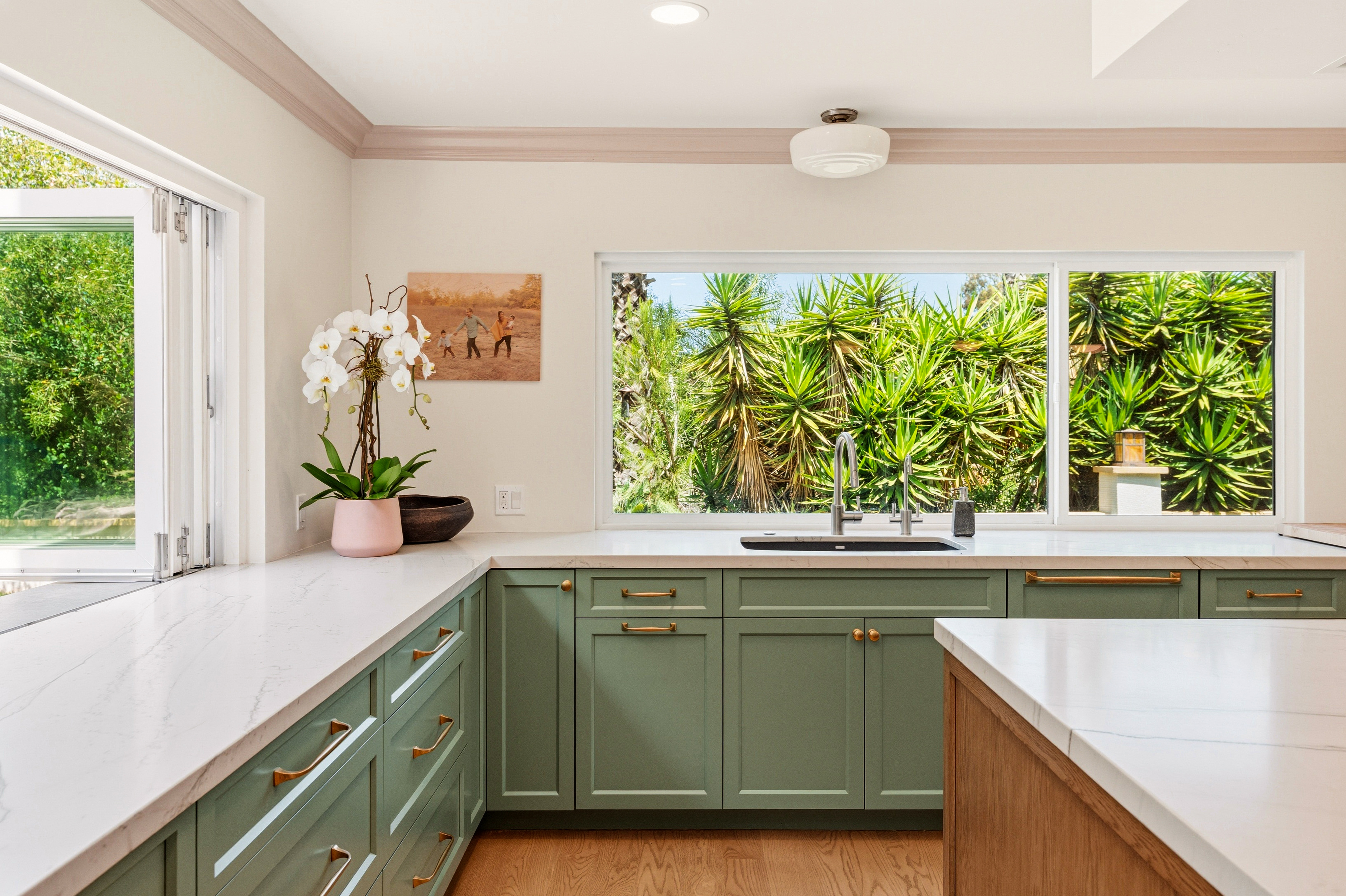Sage green sink cabinetry with brass hardware, white counters, wide garden windows and an open bi-fold window.