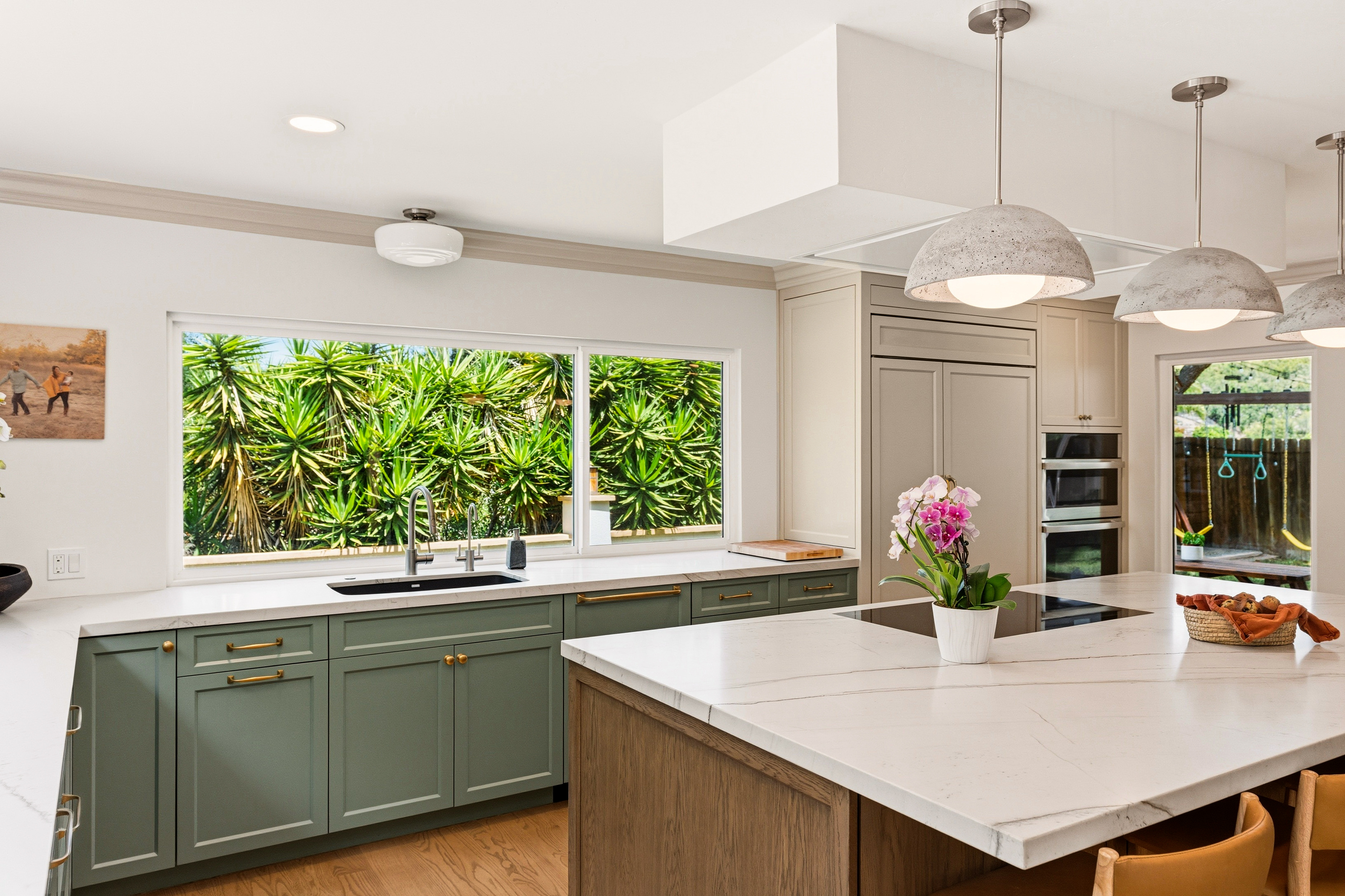 Open San Diego kitchen with sage cabinetry, wood island, white counters, pendant lights and a wide garden-facing window.