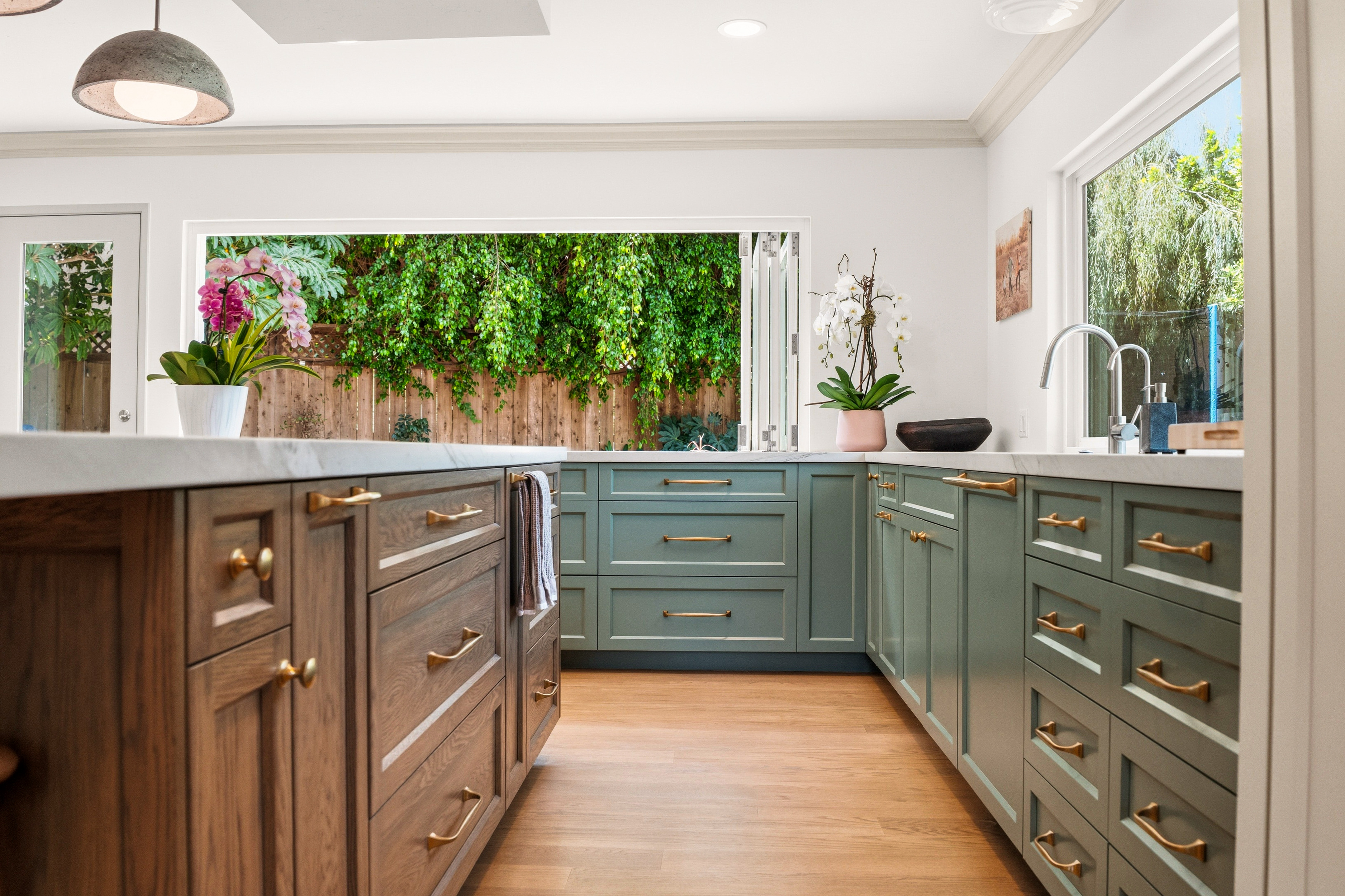 Kitchen work zone with sage cabinetry, wood island, brass hardware, white counters and an open bi-fold window.