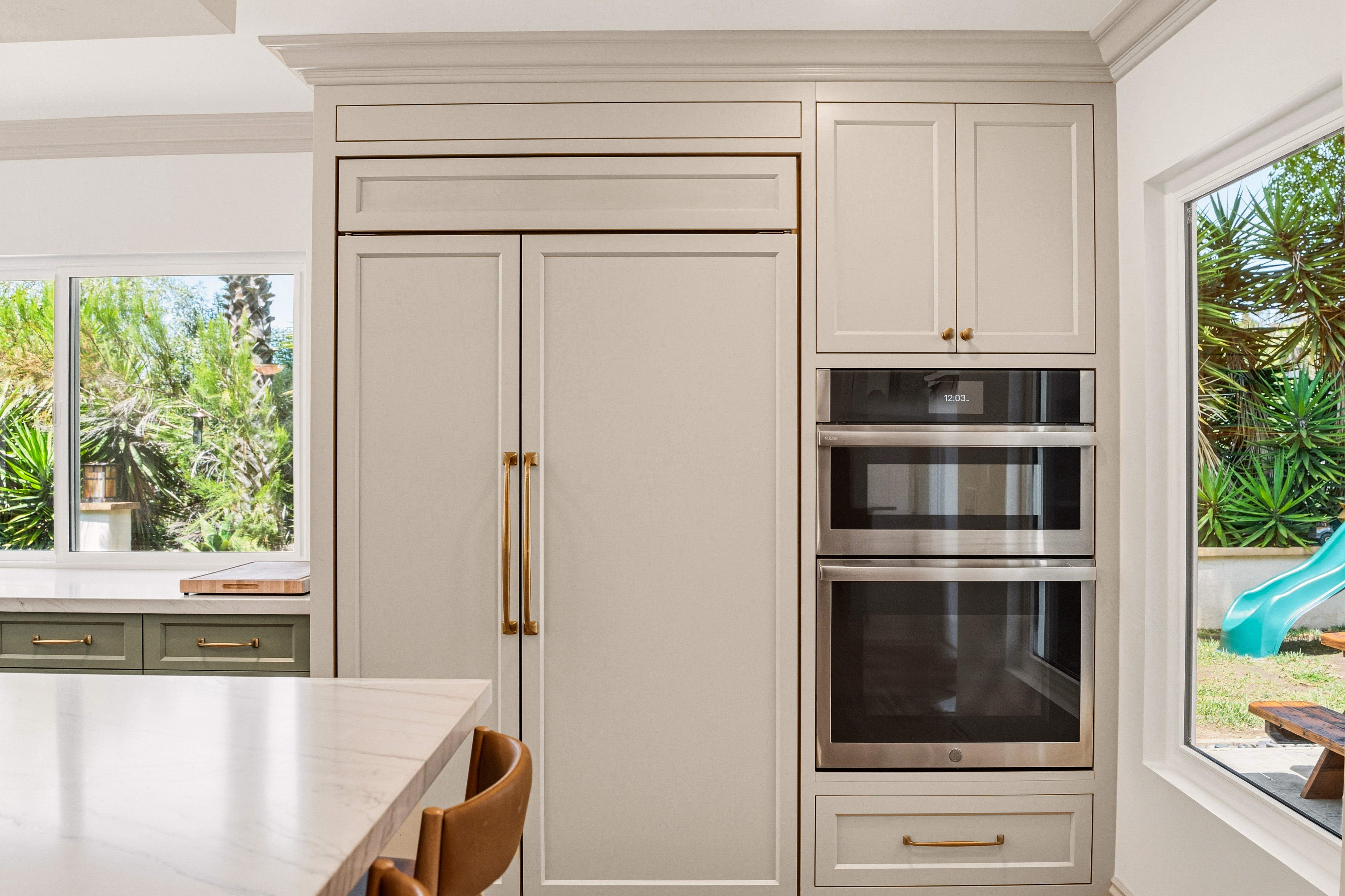 Panel-ready refrigerator and stacked stainless ovens integrated into tall white kitchen cabinetry with brass hardware.