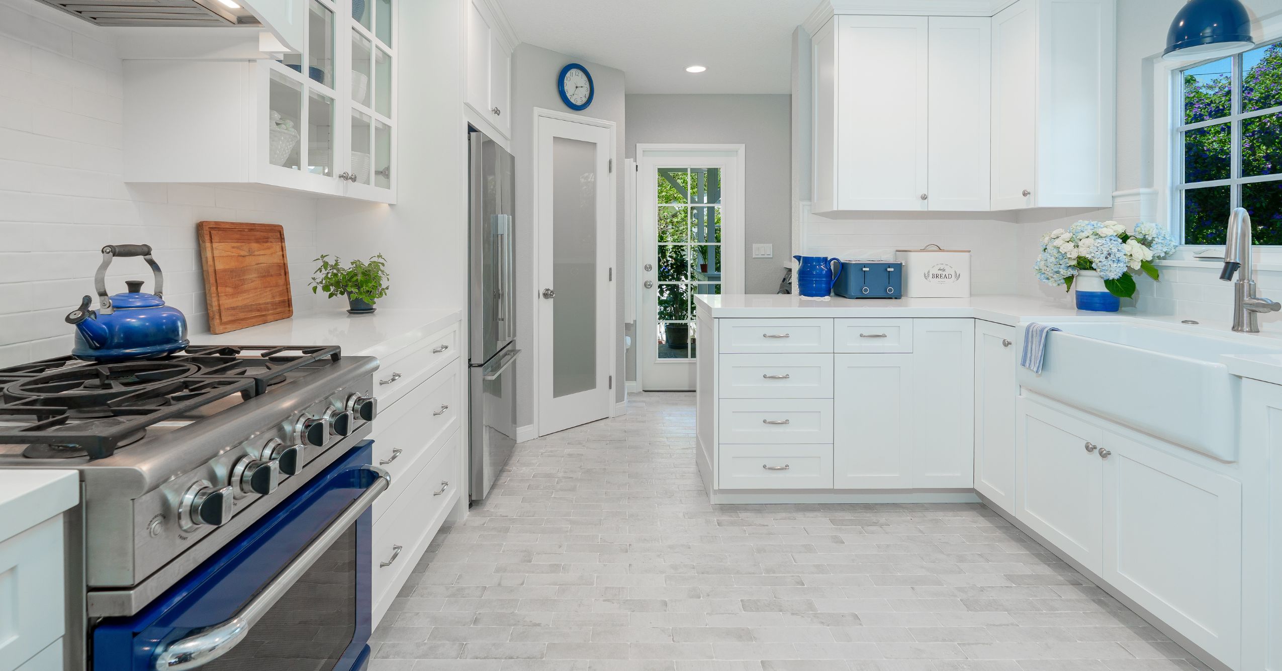 Galley-style white kitchen with blue range, stainless refrigerator, farmhouse sink, and frosted pantry door.
