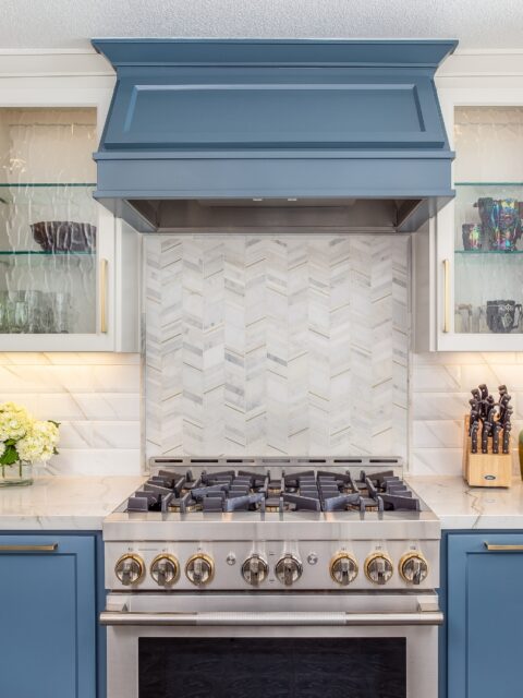 Two-tone blue cabinetry with brass hardware, chevron marble mosaic backsplash accent, glass-front white upper cabinets, quartz waterfall countertops, gas range with brass-trimmed knobs, brass gooseneck faucet, open sightline into the family room with a stacked-stone fireplace surround.