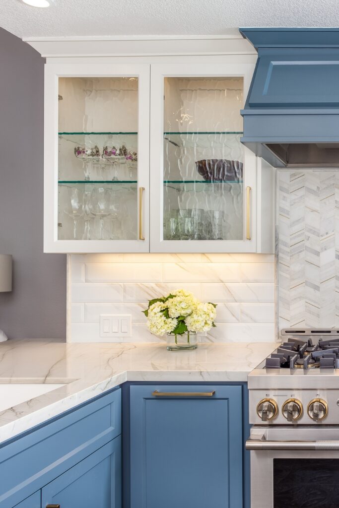 Glass-front white cabinet with glass shelving and brass pull above chevron tile backsplash