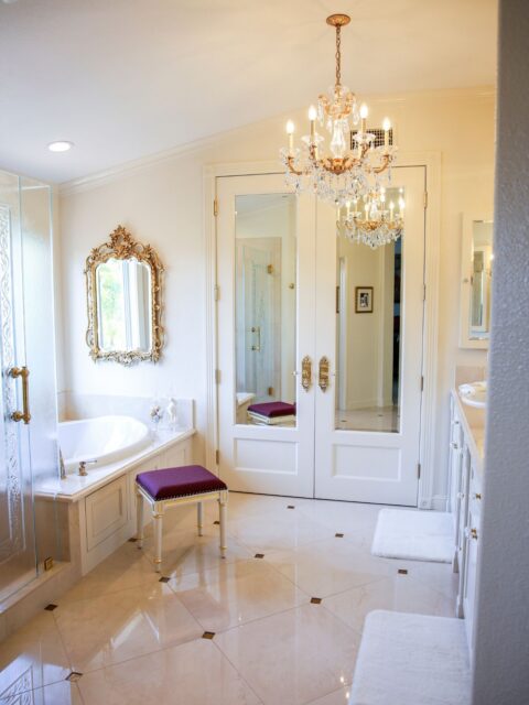 Carlsbad primary bathroom remodel with soaking tub, crystal chandelier, mirrored doors, ornate gold accents and light stone flooring.