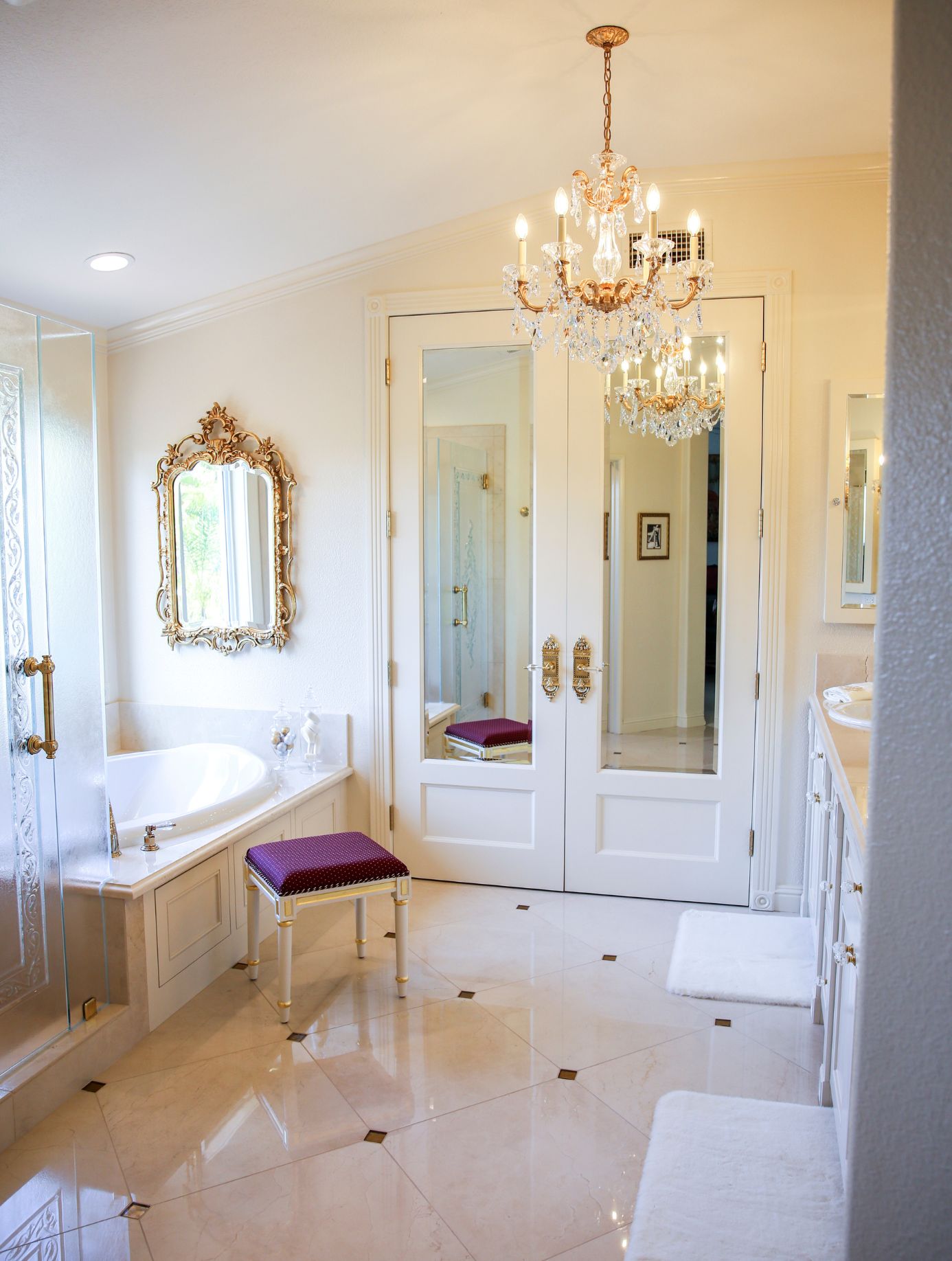Carlsbad primary bathroom remodel with soaking tub, crystal chandelier, mirrored doors, ornate gold accents and light stone flooring.