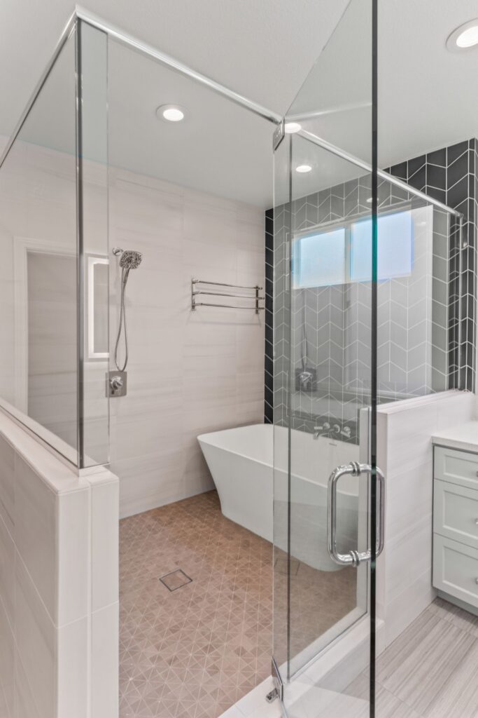 Glass-enclosed shower with handheld fixture, freestanding tub, frosted window and dark chevron accent tile.