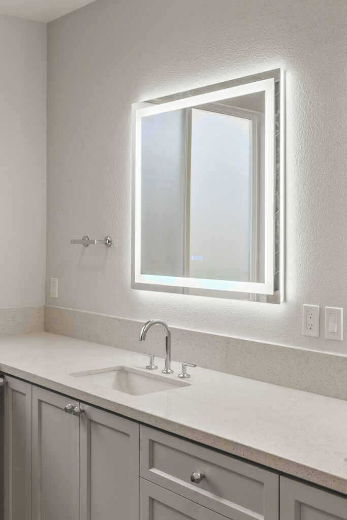 Long gray vanity with single undermount sink, illuminated mirror, chrome fixtures and speckled countertop.