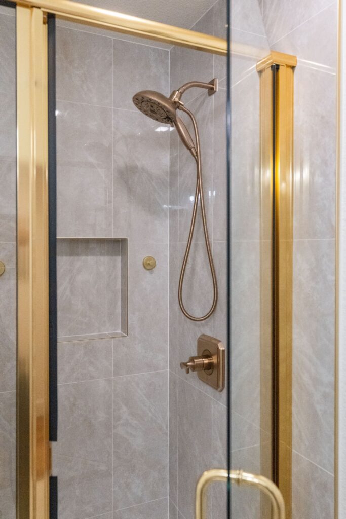 Glass-enclosed shower with gold-toned trim, large gray wall tile, niche and handheld shower fixture.