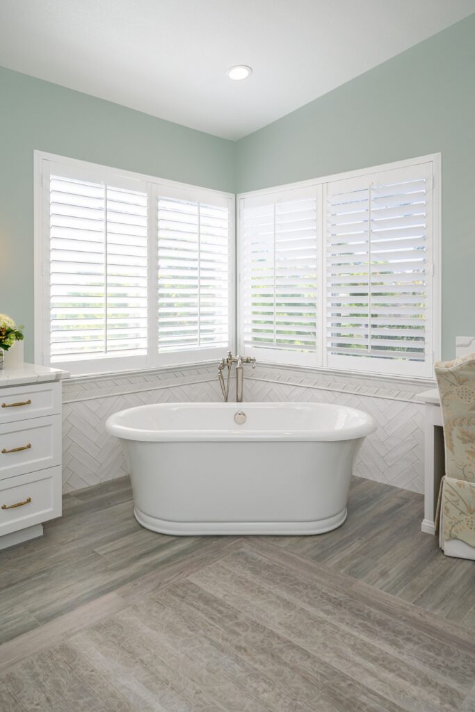 Primary bathroom with freestanding tub, white herringbone tile surround, plantation shutters and gray tile flooring.