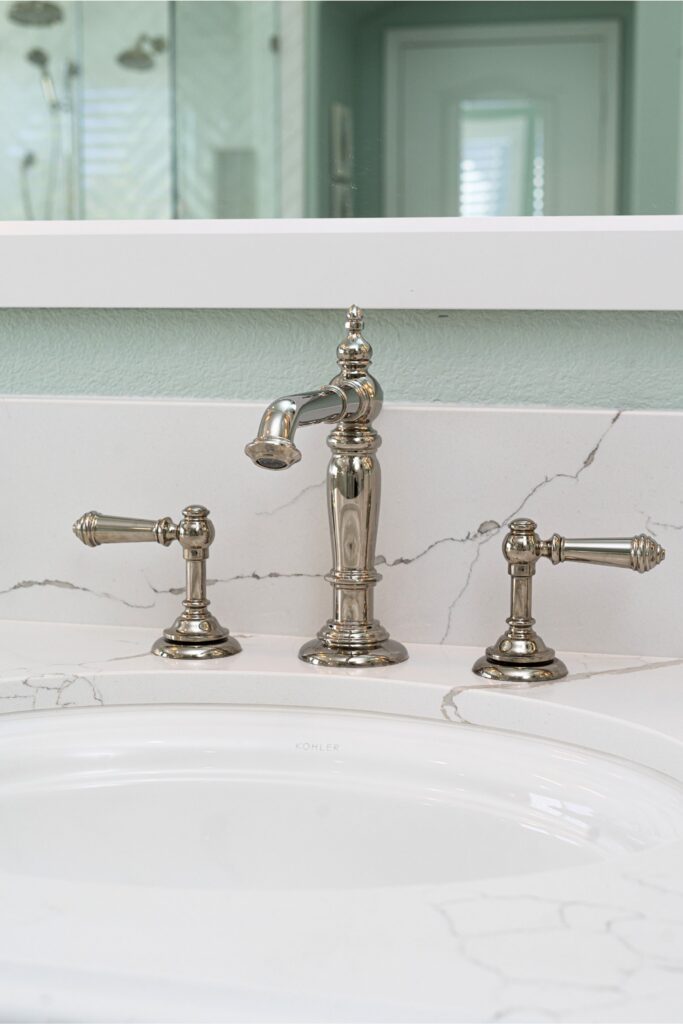 Bathroom sink with polished metal widespread faucet, veined white countertop and illuminated mirror.