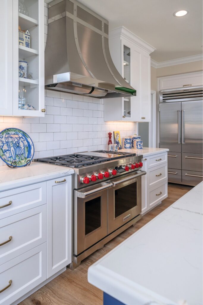 Cooking wall with stainless range, custom metal hood, white subway tile, glass-front cabinets and brass hardware.