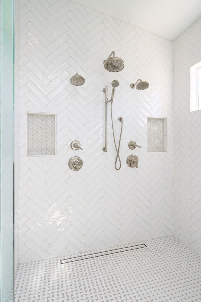 Walk-in shower with white herringbone wall tile, dual niches, multiple showerheads and mosaic tile floor.