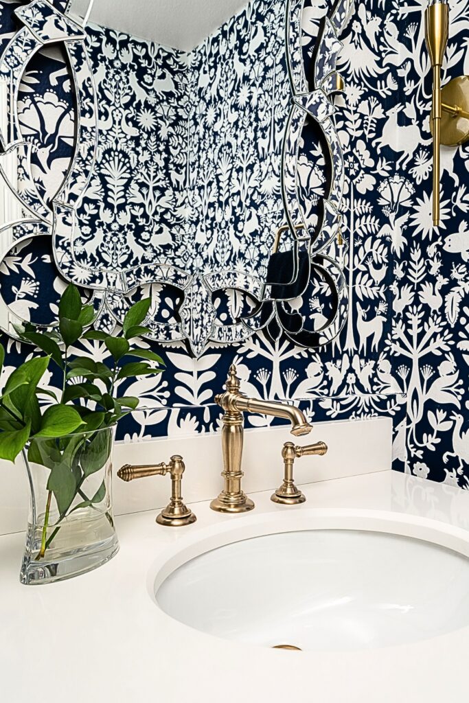 Powder room vanity with navy-and-white floral wallpaper, brass faucet, white counter and decorative mirror.
