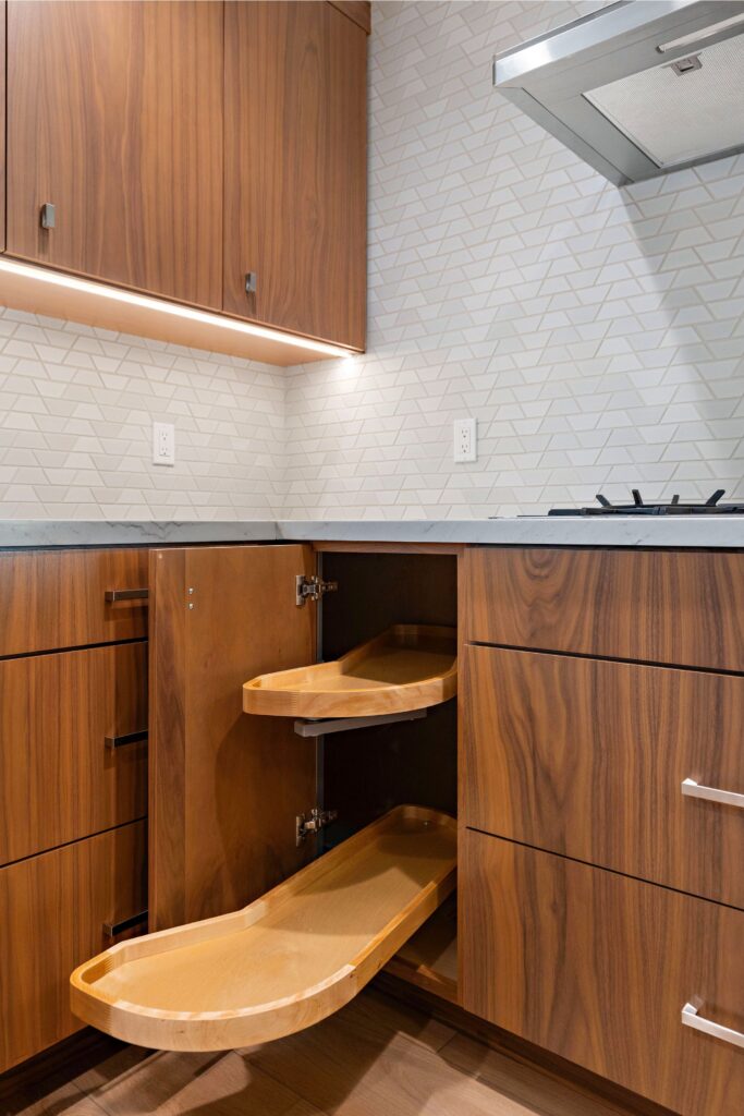 Walnut kitchen corner cabinet with pull-out wood shelves, light countertop and geometric tile backsplash.