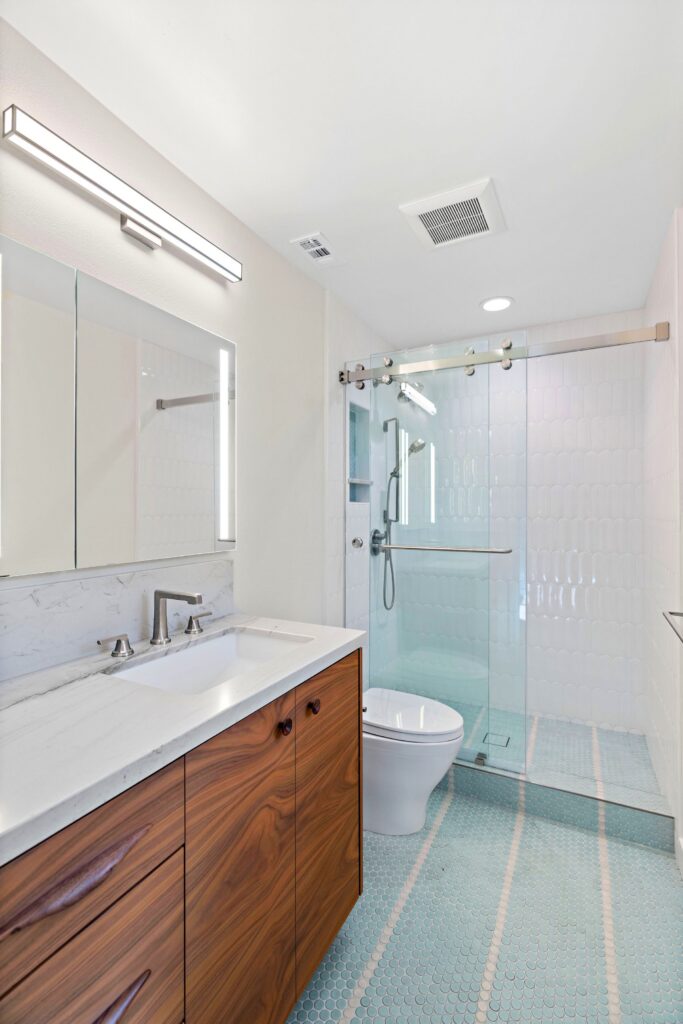 Bathroom with walnut vanity, marble-look counter, illuminated mirror, glass shower and blue penny tile floor.
