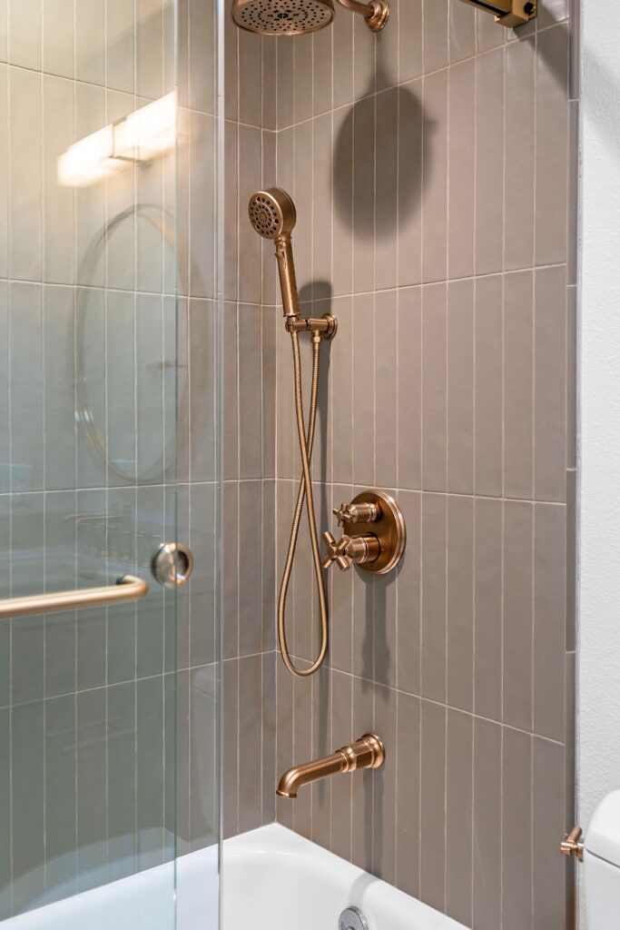 Tub and shower with vertical gray tile, bronze rain shower, handheld fixture and glass enclosure.
