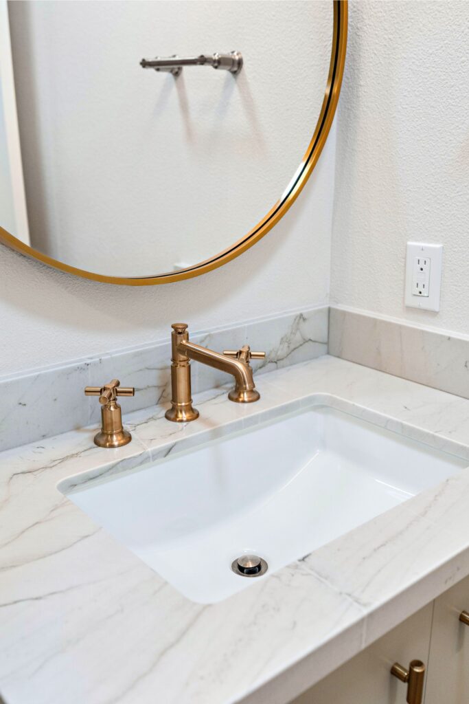Bathroom vanity with undermount sink, marble-look countertop, bronze faucet and round brass-framed mirror.