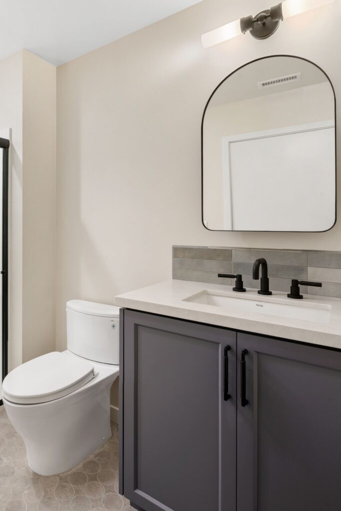 Guest bath with gray vanity, white counter, black fixtures, arched mirror and gray tile backsplash.