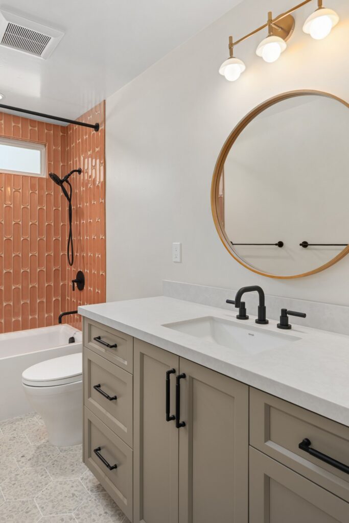 Bathroom with gray vanity, black fixtures, round mirror, coral shower tile and patterned floor tile.