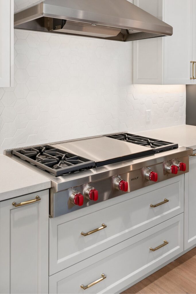 Kitchen cooking wall with stainless range, vent hood, white cabinetry, brass pulls and textured white hex backsplash.