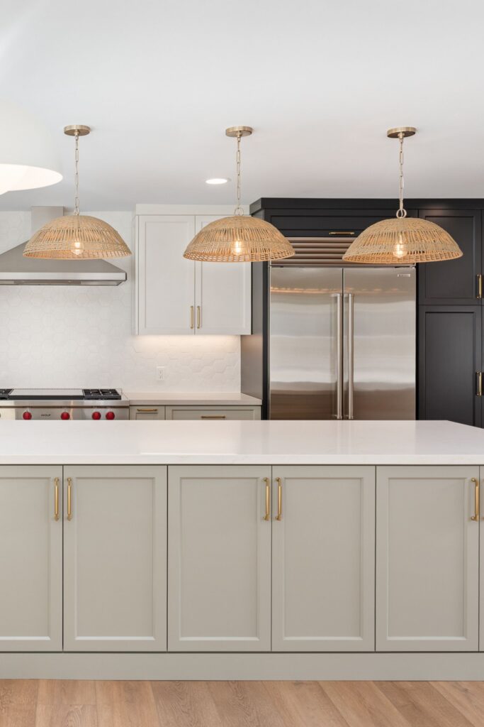 Kitchen island with light cabinetry, white countertop, woven pendant lights, stainless refrigerator and dark pantry cabinets.