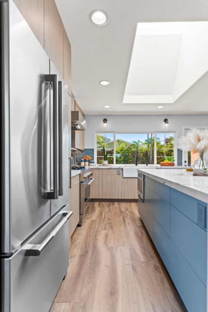 Kitchen walkway with stainless refrigerator, blue island, light wood cabinetry and large ceiling skylight