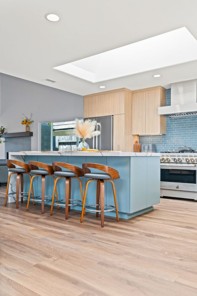 Blue kitchen island with waterfall stone countertop, wood bar stools, light cabinetry and skylight