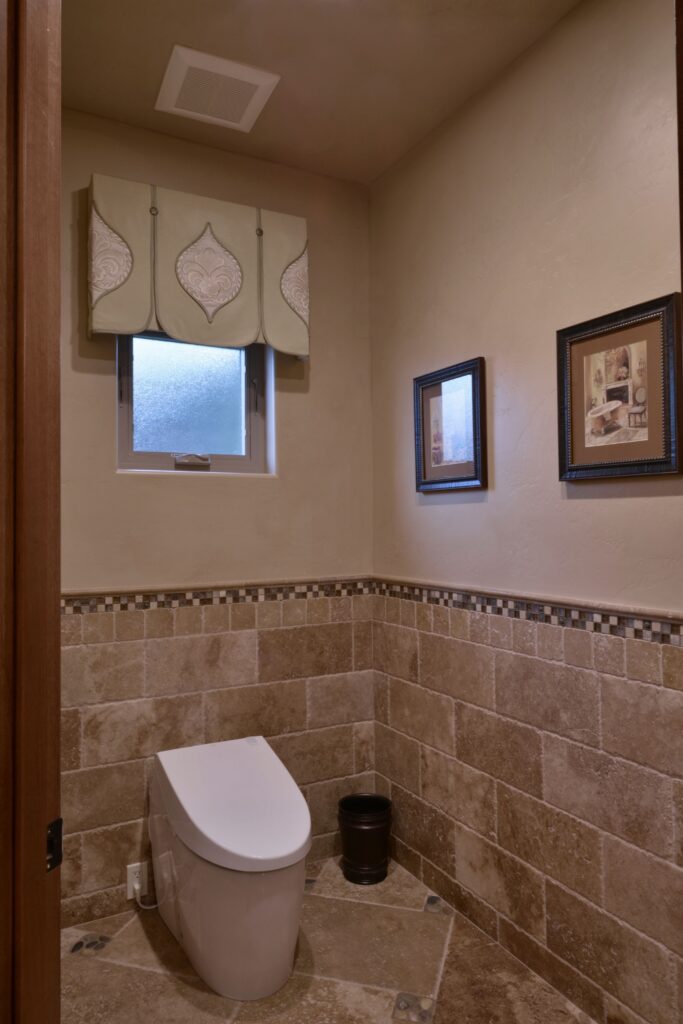 Toilet alcove with stone tile wainscoting, mosaic border, tiled floor and frosted window with fabric valance.