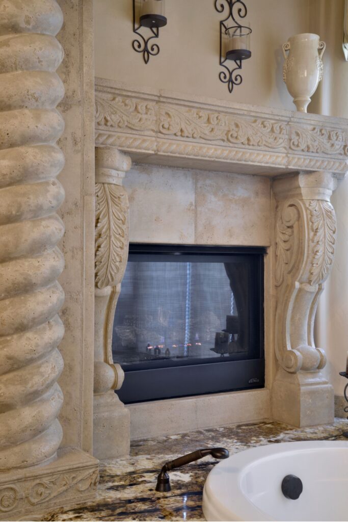 Carved stone fireplace beside the bathtub with granite deck, decorative mantel and sculpted architectural detailing.