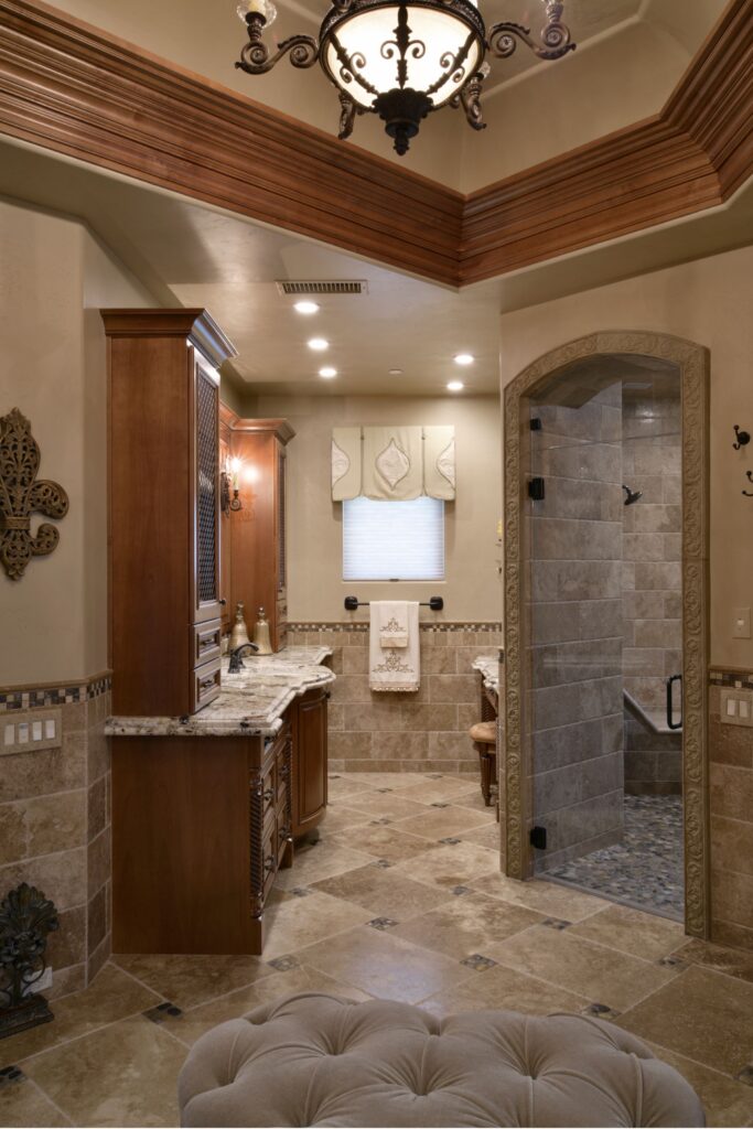 Primary bathroom with wood vanities, granite counters, stone tile flooring, arched glass shower and chandelier.