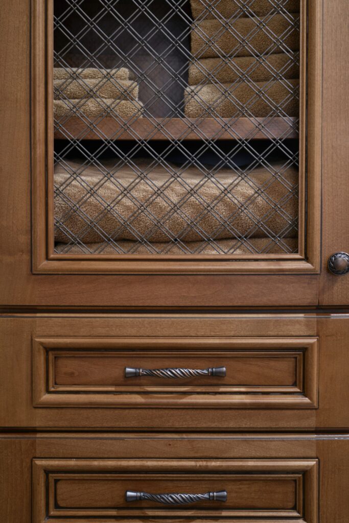 Custom wood bathroom storage cabinet with wire-mesh door, towel shelving, paneled drawers and dark metal hardware.