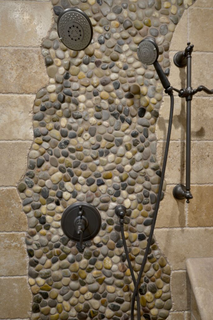 Shower wall with pebble mosaic accent, dark metal rain shower, handheld sprayer and control fixtures.