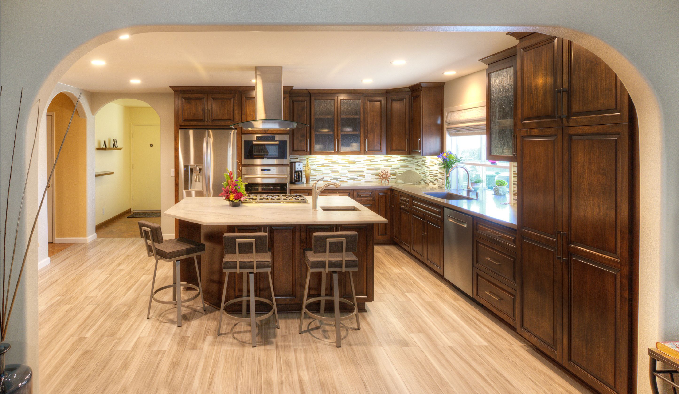 Wide Mira Mesa kitchen view with arched opening, dark wood cabinets, central island, mosaic backsplash and stainless appliances.