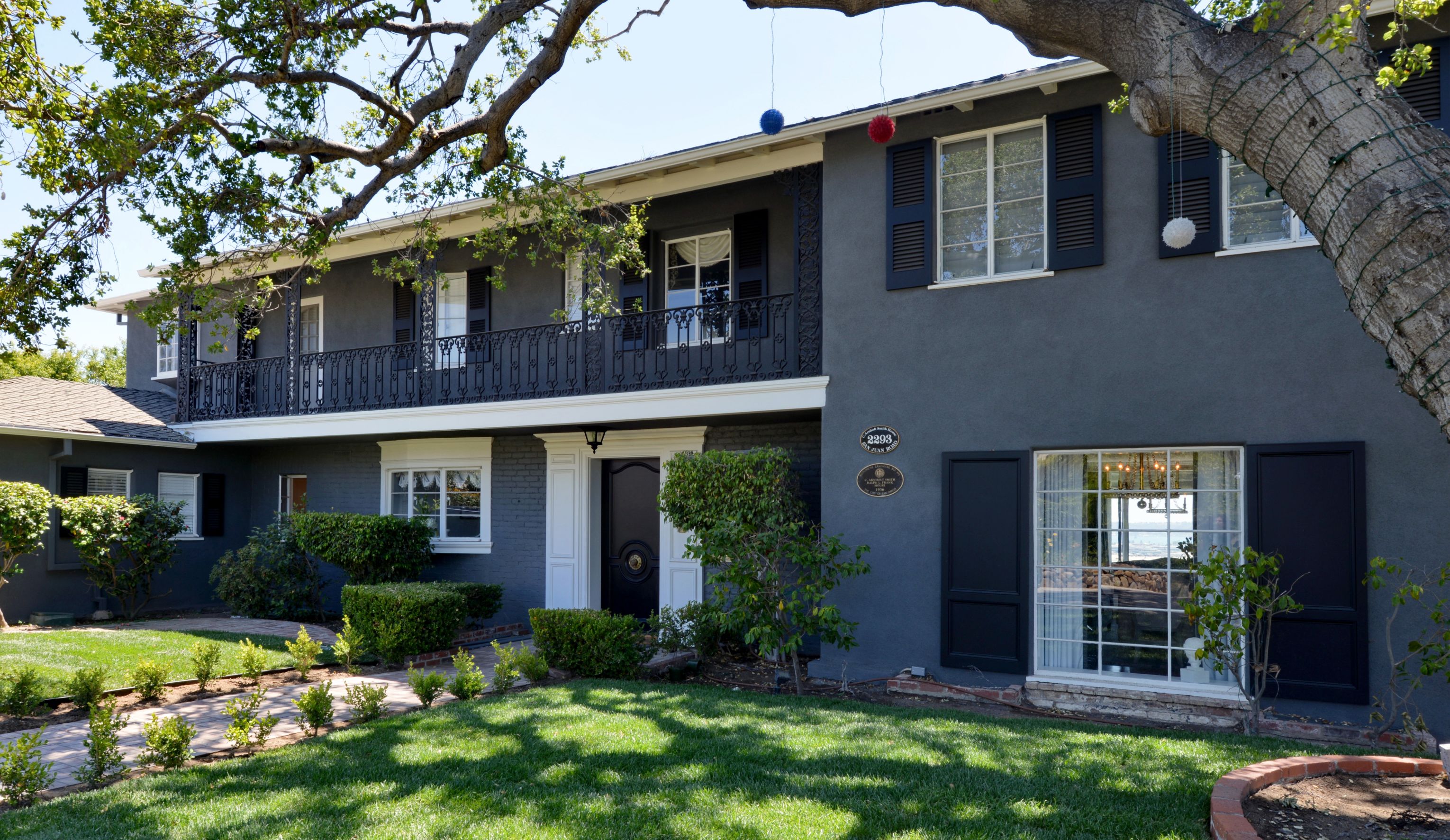 Mission Hills remodeled home exterior with dark stucco, black shutters, upper balcony, mature trees and landscaped lawn.