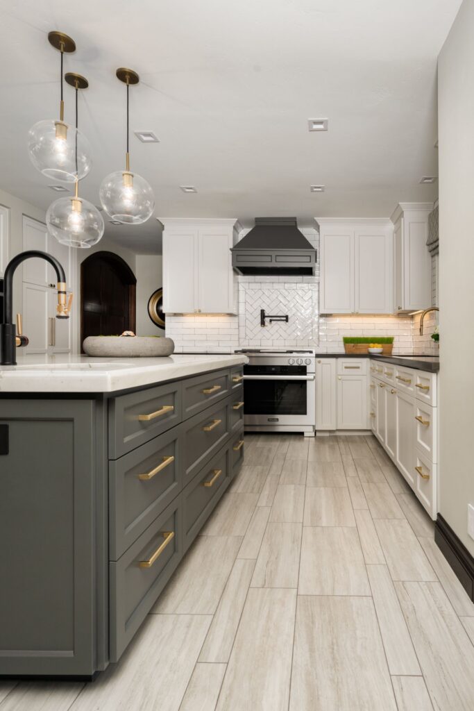 Kitchen with gray island, white cabinetry, brass hardware, glass pendants, dark range hood and light tile flooring