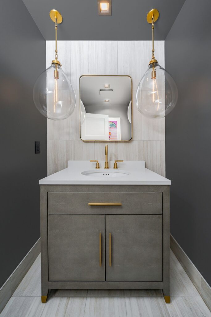 Powder room with gray vanity, white countertop, brass fixtures, framed mirror and glass pendant lights
