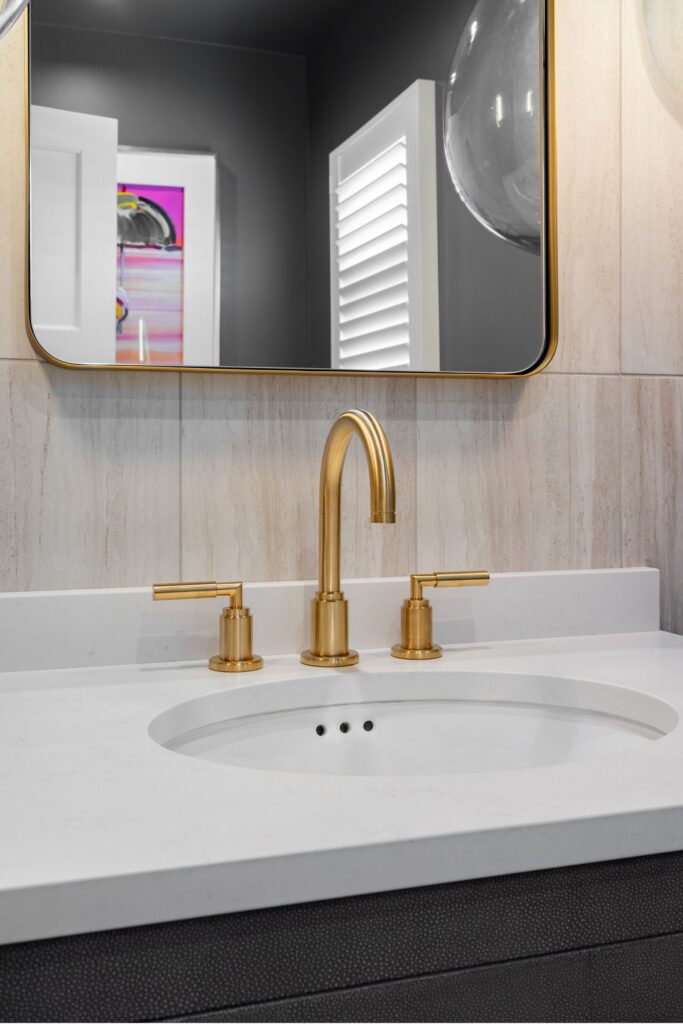 Powder room sink with brass widespread faucet, white countertop, brass-framed mirror and vertical wall tile