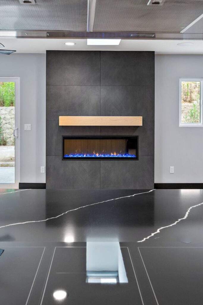 Living room fireplace with dark slab surround and wood mantel viewed across a black veined kitchen island