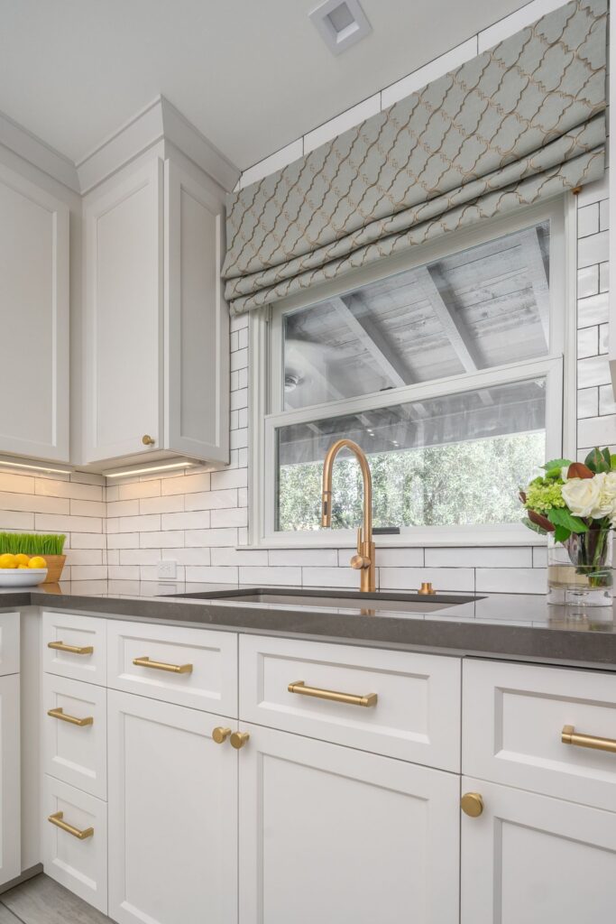 Kitchen sink wall with white shaker cabinets, brass faucet, dark countertop, subway tile backsplash and window