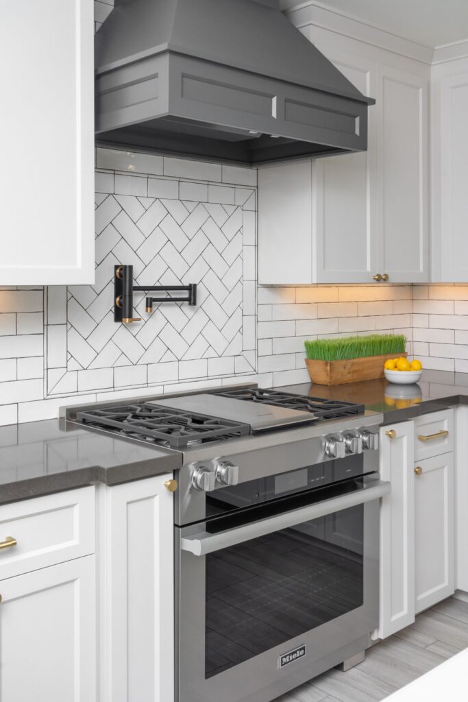 Stainless range beneath dark gray hood with herringbone tile backsplash, white cabinetry and brass hardware