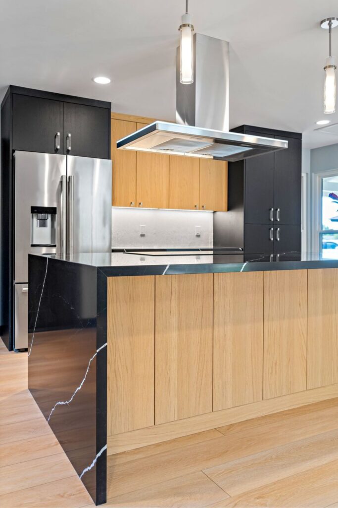 Kitchen island with black veined waterfall counter, wood cabinetry, stainless hood and pendant lighting