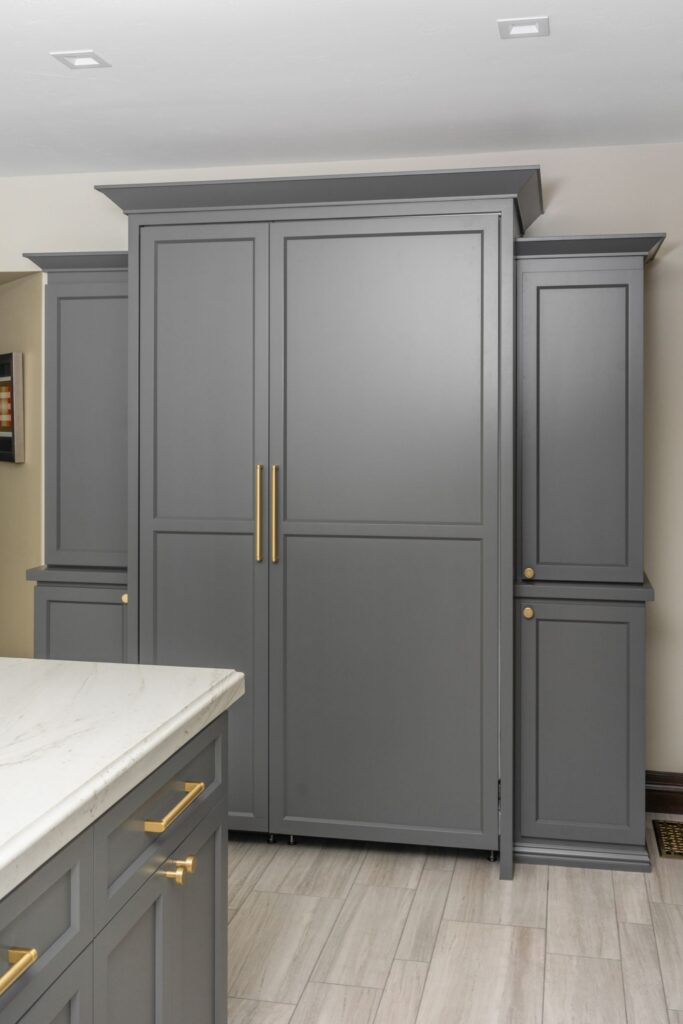 Tall gray kitchen cabinetry with brass pulls beside matching island and light rectangular tile flooring
