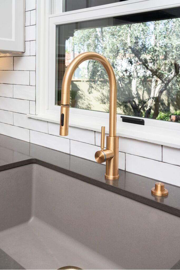 Brass kitchen faucet over gray undermount sink with dark counter, white subway tile backsplash and window