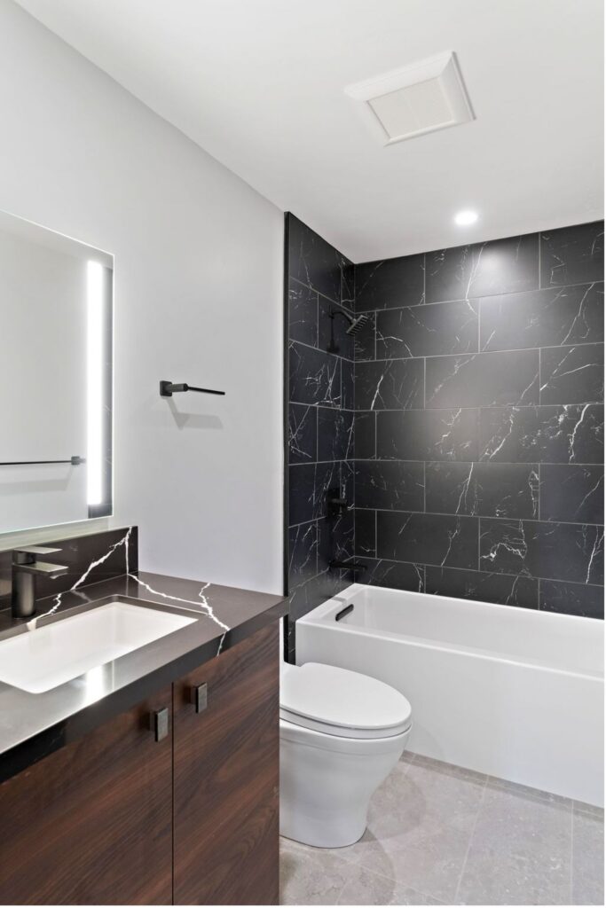 Bathroom with dark wood vanity, black veined countertop, illuminated mirror and black tile tub surround