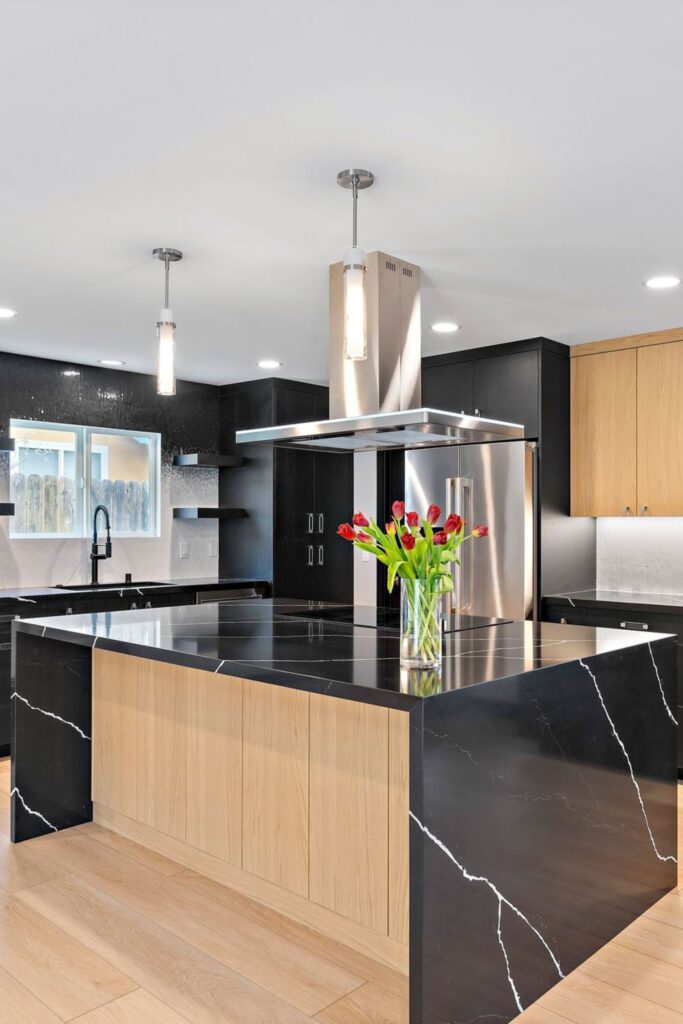 Kitchen island with wood cabinetry, black waterfall countertop, stainless hood and pendant lights