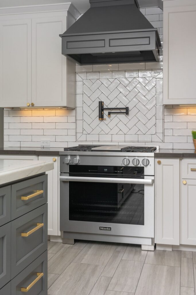 Kitchen range wall with stainless range, gray hood, white herringbone tile and brass-accented cabinetry